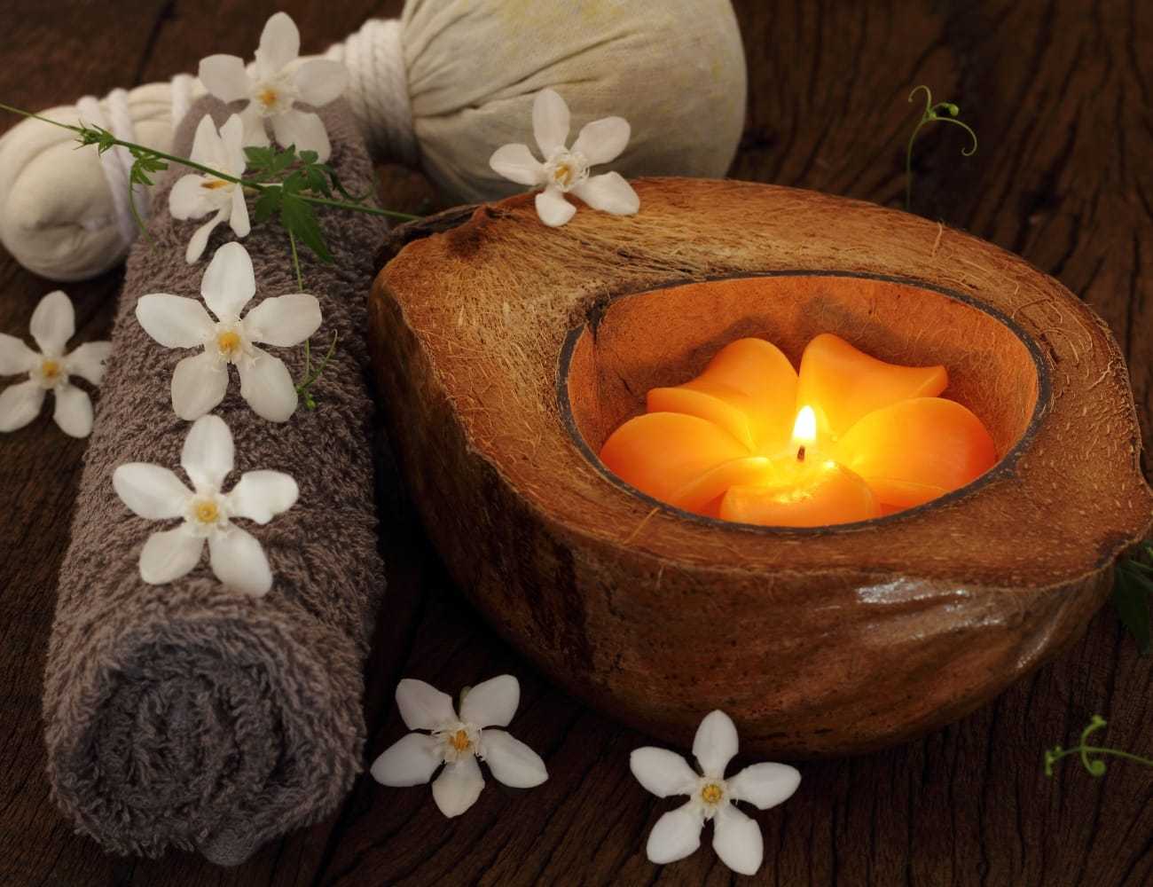 Candle in a coconut shell with flowers and a rolled towel on a wooden surface.