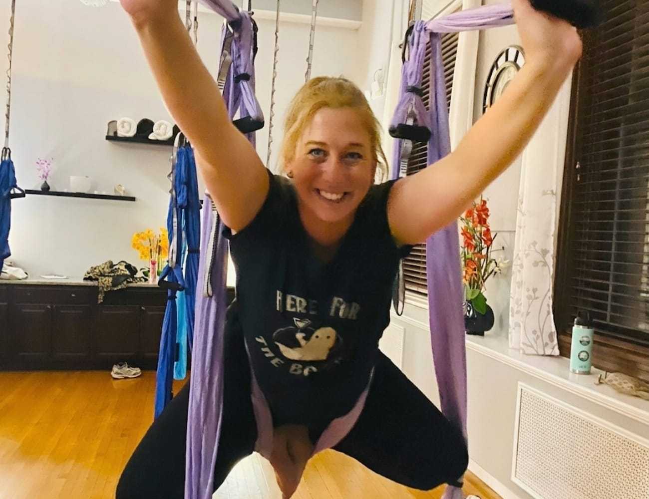 A woman smiling while doing aerial yoga in a room with purple hammocks.