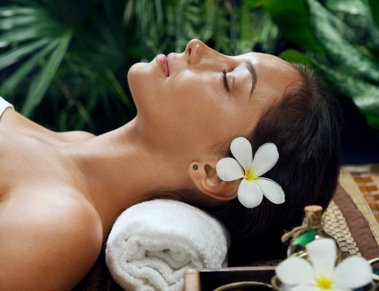 Woman relaxing with flower in hair, lying on a rolled towel amidst lush greenery.